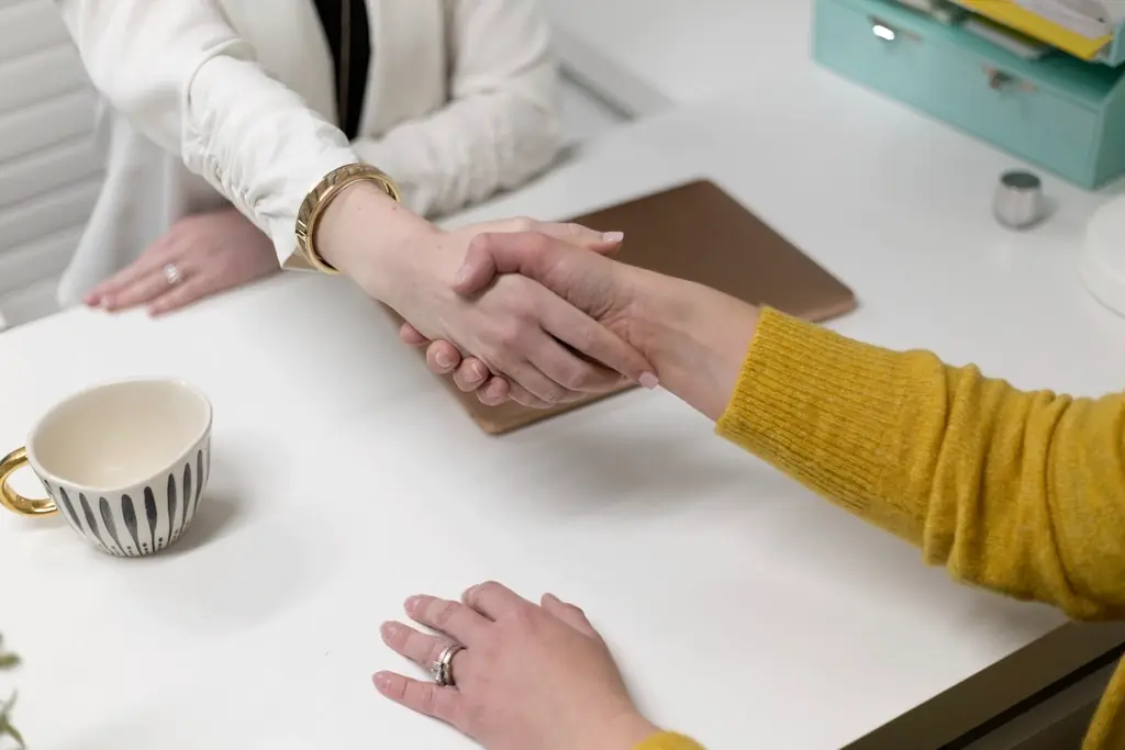 Two people shaking hands across a desk at the close of an interview